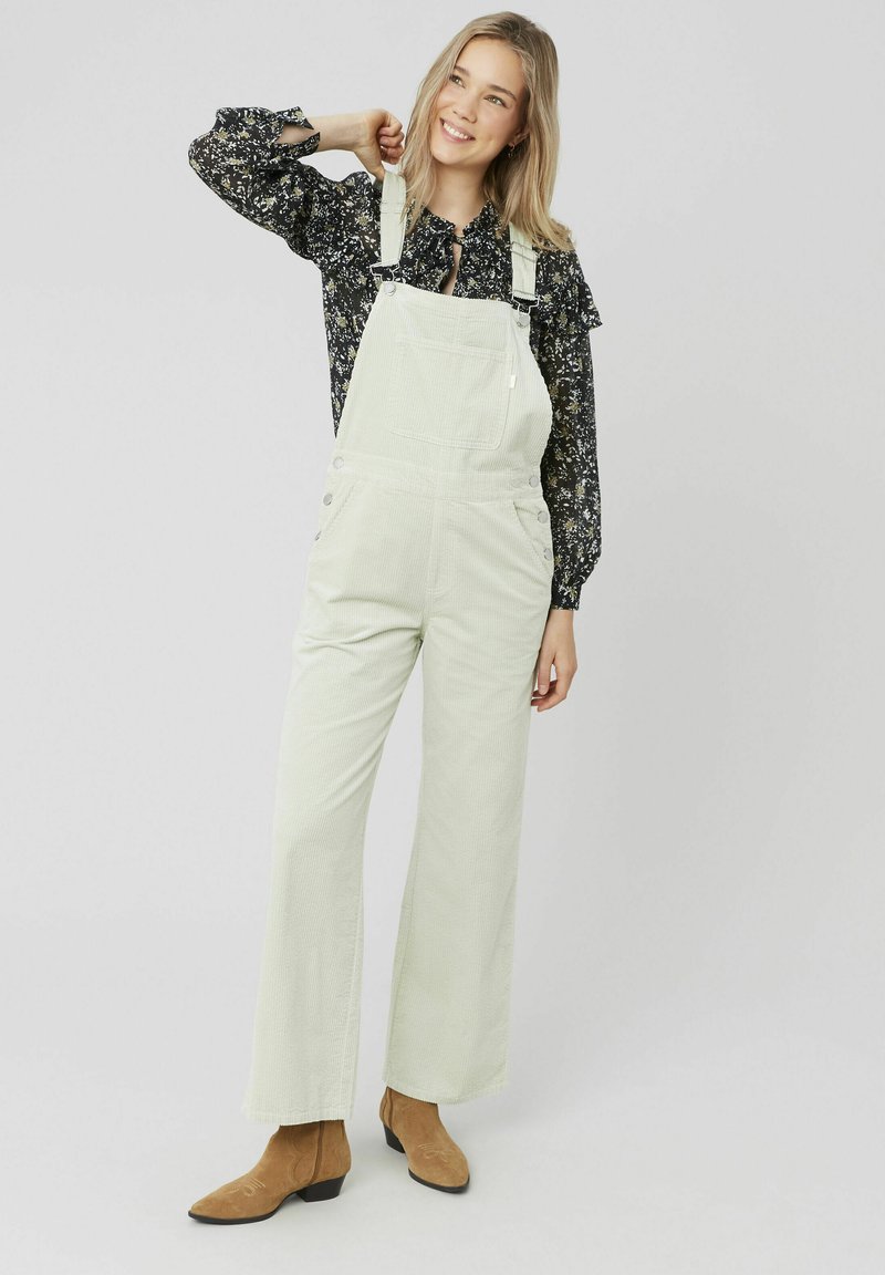 Salopette en velours côtelé vert clair avec des jambes larges et une poche avant, portée par-dessus un blouse à manches longues noire à fleurs. Des bottines brunes à la cheville complètent le look.