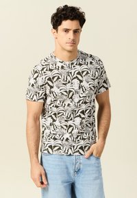 Camiseta de manga corta con base verde y un estampado de hojas tropicales blancas, cuello redondo y corte relajado, combinada con pantalones vaqueros azules claros.