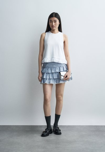 Jeune femme aux longs cheveux noirs portant un haut blanc sans manches, une mini-jupe à volants bleue superposée, des chaussettes noires, des mocassins, tenant une pochette argentée contre un mur gris.