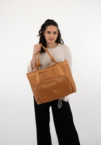 Beżowa torba na ramię wykonana z teksturowanego materiału z dwoma węzłowymi uchwytami, siatkową przednią kieszenią oraz tłoczonym logo wzdłuż górnej krawędzi.