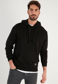 Felpa nera con tasca frontale, cappuccio con cordino, polsini a coste e fondo a coste. Indossata sopra una maglietta bianca. Design semplice e casual.