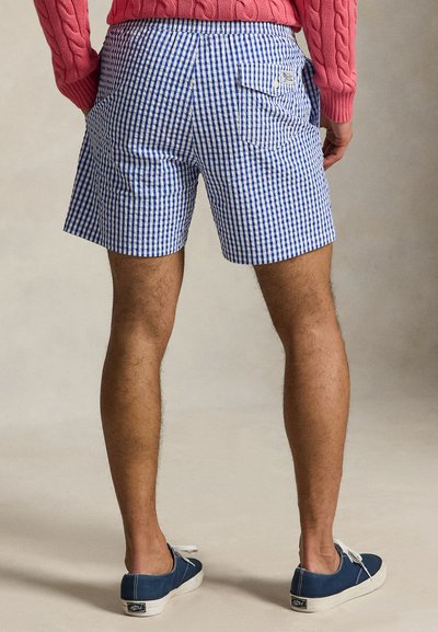 Homme portant un short à carreaux bleu et blanc, un pull en maille torsadée rose et des baskets bleu marine, debout sur un fond neutre.