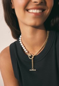 Zlatý retiazkový náhrdelník so hviezdnym akcentom, obsahujúci časť veľkých perál. Materiály zahŕňajú kov a perly; minimalistický dizajn, elegantný.