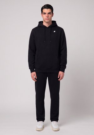 Giovane uomo in piedi, indossa una felpa con cappuccio nera con un piccolo logo bianco, pantaloni neri e scarpe da ginnastica bianche su uno sfondo chiaro e uniforme.