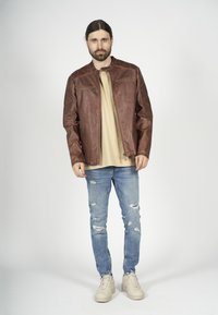 Brun läderjacka med kviltade axlar, framdragkedja, två dragkedjefickor, beige skjorta under, slitna blå jeans och ljusa sneakers.