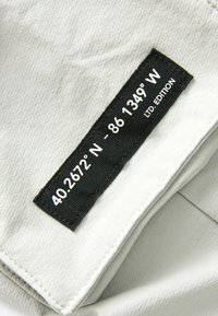 Tissu gris clair avec une surface texturée ; présente une étiquette en tissu noir indiquant les coordonnées et "ÉDITION LIMITÉE."
