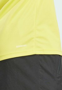 Yellow mesh athletic shirt with short sleeves, featuring a breathable texture and "AEROREADY" branding at the hem. Black pants partially visible.