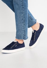 Baskets en toile bleu marine avec semelles en caoutchouc blanches et œillets dorés, associées à un jean bleu déchiré coupé aux chevilles.