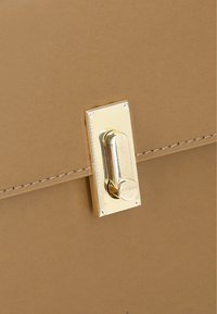 Detail van een tan lederen tas met een gouden rechthoekige sluiting met een ronde handgreep en contrasterende stiksels langs de randen.