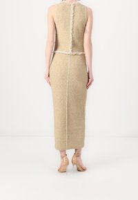 Robe beige sans manches en tissu texturé, bords effilochés et couture au dos. Portée avec des chaussures à talons transparents.