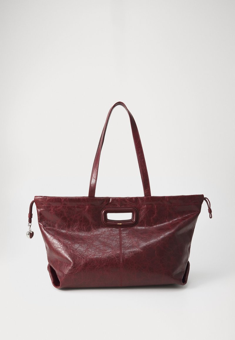 Crimson leather tote bag with a textured finish, dual handles, and a prominent front opening. Features a decorative silver charm on the side.