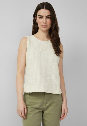 Vrouw met lang donker haar, gekleed in een mouwloos, gestructureerd wit topje en olijfgroene broek, staand tegen een effen grijze achtergrond.