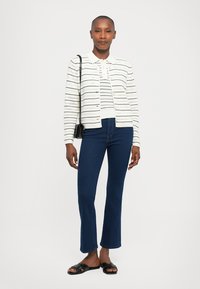 Witte gebreide cardigan met horizontale strepen, gecombineerd met donkerblauwe flared jeans en zwarte instappers. Eenvoudig ontwerp, lichte stof.