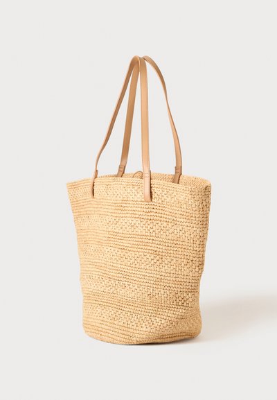 Sac cabas en paille tissée beige avec deux longues bandoulières en cuir marron clair sur un fond clair uni.