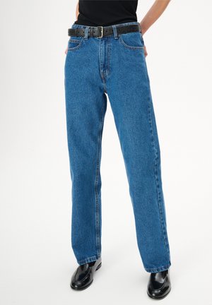 Osoba ubrana w niebieskie, proste dżinsy z wysokim stanem, czarny pasek, czarny top i czarne skórzane buty na białym tle.