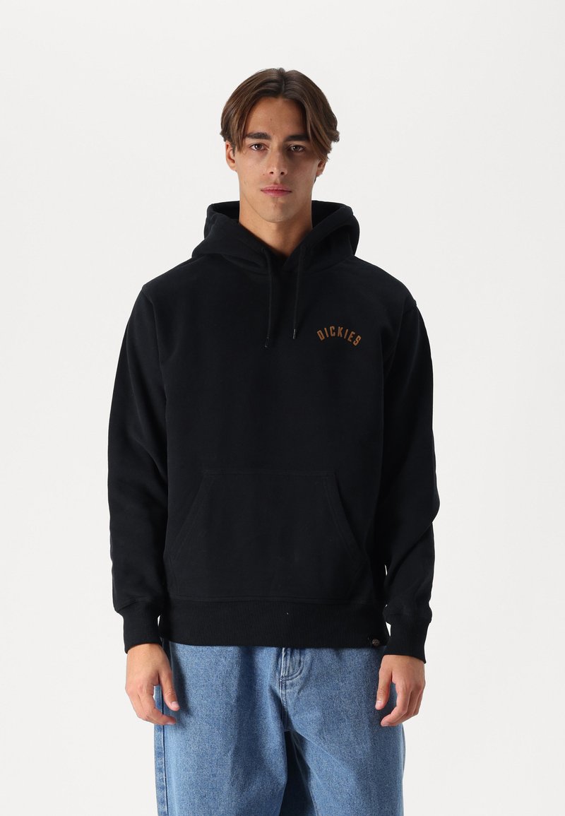 Sudadera negra con un bolsillo tipo canguro, capucha con cordón y un pequeño logo marrón en el pecho. Hecha de un tejido texturizado, combinada con jeans azules.
