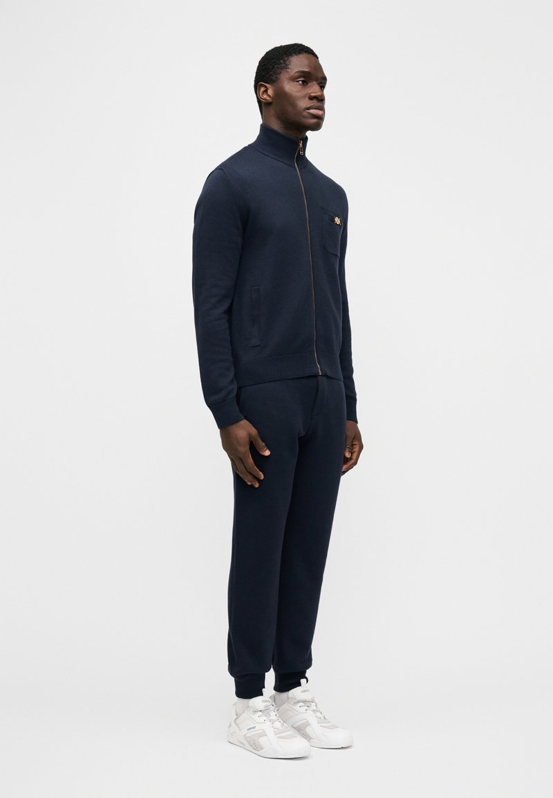 Homme debout de profil gauche, portant un pull à fermeture éclair bleu marine avec poche sur la poitrine, un pantalon de jogging assorti et des baskets blanches, sur un fond uni.