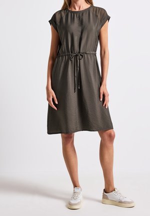 Femme portant une robe olive foncé mi-longue à manches courtes avec une taille resserrée par un cordon et des baskets blanches décontractées, debout sur un fond uni.