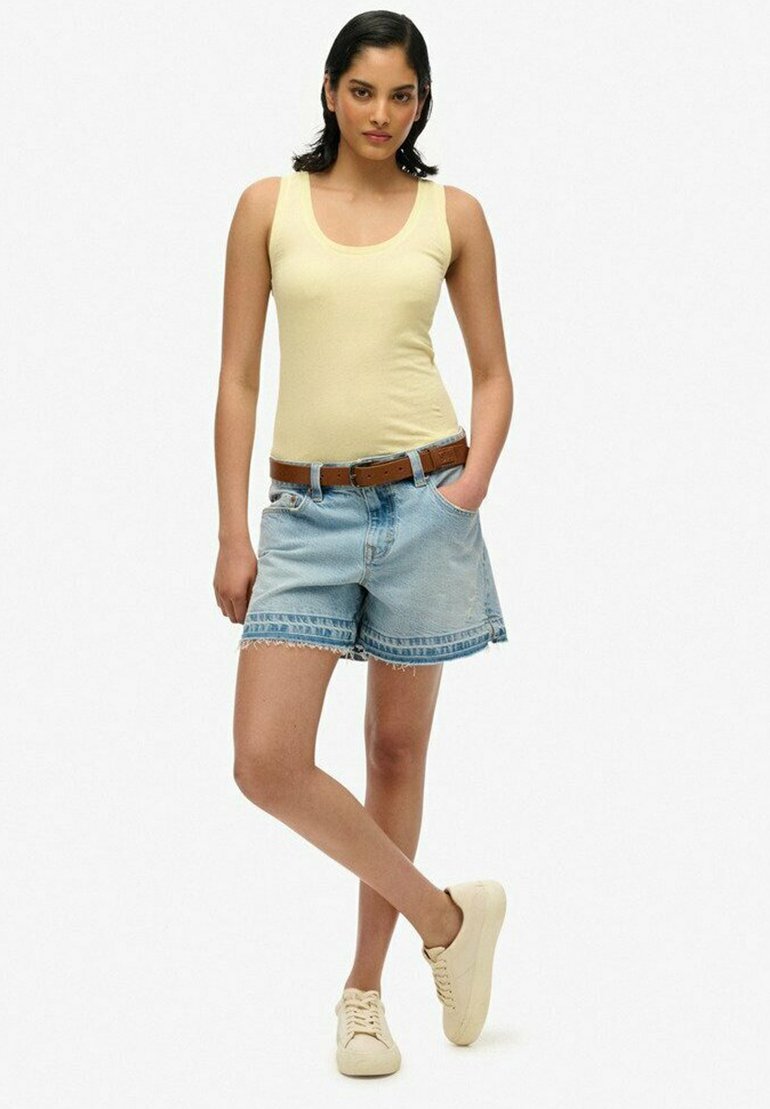Lichtgele tanktop, blauwe denimshorts met franjes, bruine riem en beige sneakers. Model staat in een ontspannen houding voor een witte achtergrond.