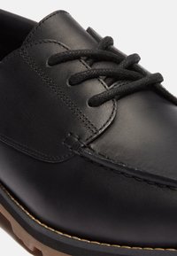 Zapato de cuero negro con una textura suave, punta redonda y cordones negros gruesos. Presenta detalles de costura y una suela de color marrón claro que contrasta.