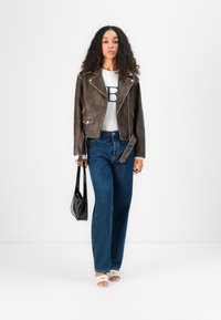 Brun lærbikerjakke med sølvdetaljer, hvit grafisk t-skjorte, vide blå jeans, svart håndveske og beige sandalferd.