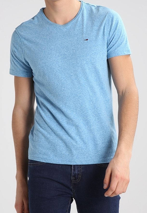 Homme portant un t-shirt bleu clair à manches courtes et un jean bleu foncé, debout devant un fond clair uni.