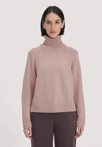 Rosa gestrickter Pullover mit hohem Rollkragen, gerippten Bündchen und Saum, mit lockerer Passform und strukturiertem Stoff. Kombiniert mit dunklen Hosen.