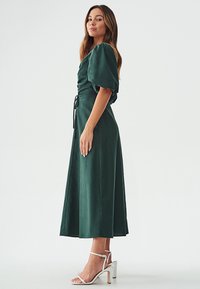 Green, mid-length dress featuring puff sleeves, tied waist, and soft texture, complemented by white, strappy high-heeled sandals.
