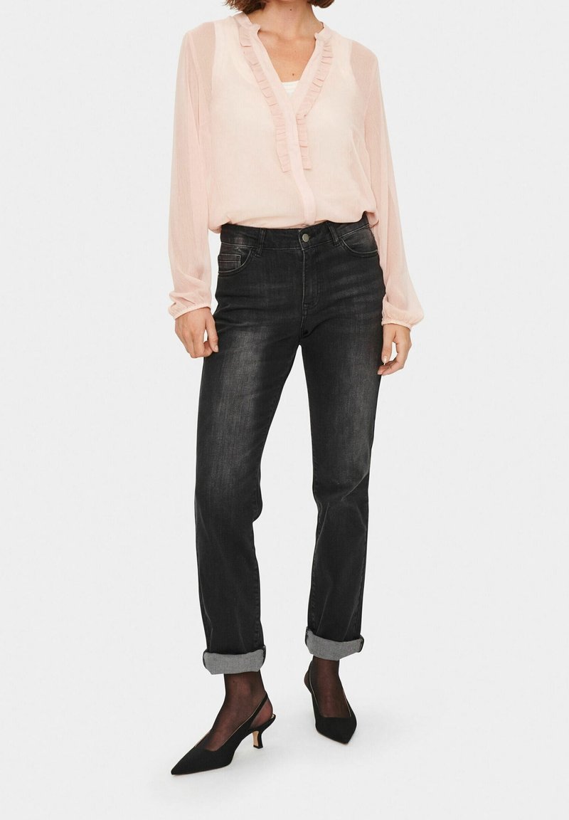 Helle rosa, rüschige Bluse mit langen Ärmeln, kombiniert mit dunkelblauen Jeans, hochgekrempelten Säumen und schwarzen spitzen Pumps mit niedrigem Absatz.