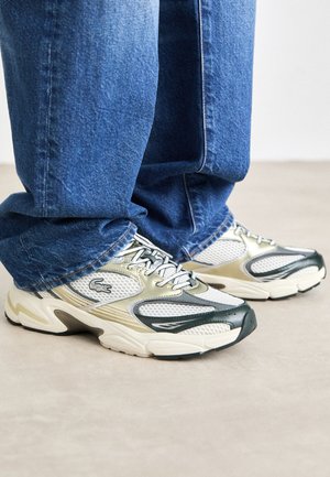 Sneakers featuring a mesh upper with gold and dark green accents, rounded toe, thick sole, and lacing system, paired with blue denim jeans.