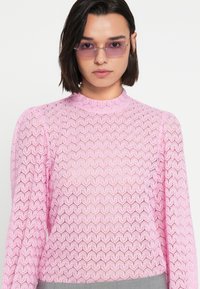 Ροζ πλεκτό τοπ με υφή σε σχήμα ζιγκ-ζαγκ, μακριά μανίκια και ψηλό λαιμό. Διαφανές υλικό με εφαρμοστή σιλουέτα.