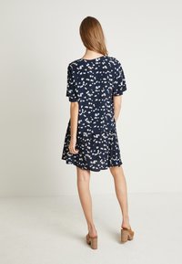 Abito navy con stampa floreale bianca, maniche corte, vestibilità relaxed, orlo a strati con volant, realizzato in tessuto morbido, indossato con sandali con tacco a blocco color tortora.