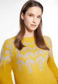Maglione giallo lavorato a maglia con motivo a foglia in grigio chiaro, caratterizzato da un colletto rotondo e maniche lunghe, mostrato in un'inquadratura ravvicinata.