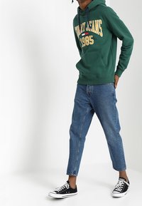 Zielona bluza z kapturem z napisem "Tommy Jeans 1985", zestawiona z jasnoniebieskimi zwężanymi dżinsami i czarnymi trampkami z białą podeszwą. Gładki materiał i swobodny krój.