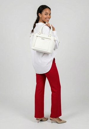 Donna sorridente con capelli intrecciati che indossa una camicetta bianca, pantaloni rossi e tacchi beige, con una borsa bianca a tracolla su uno sfondo grigio.
