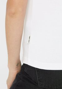 White cotton t-shirt with short sleeves, featuring a small label on the side. Paired with black denim, showing a casual outfit detail.