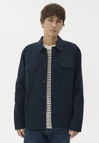 Moški nosi mornarsko zapončeno jakno preko bele in mornarsko prugaste srajce, stoji ob belem ozadju.