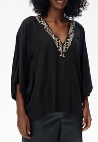 Schwarze Tunika mit V-Ausschnitt, die eine aufwendige beige Stickerei entlang des Ausschnitts aufweist. Lockere Passform mit weiten Ärmeln. Glatte Textur.