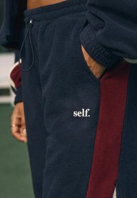 Tumesinised fliis-püksid maroonide külgede paneelidega, elastse vöökoha ja küljetaskutega. Ees on väike valge logo tekstiga "self.".