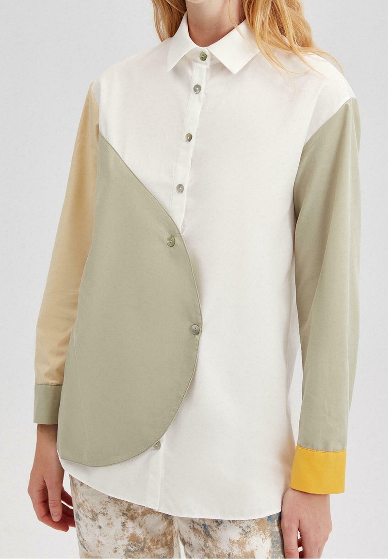 Femme porte une chemise boutonnée avec des panneaux asymétriques beige et olive, une manche beige, une manche olive avec un poignet jaune, et un pantalon à motifs.