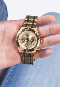 Montre en acier inoxydable doré avec un cadran rond, un cadran doré clair, des accents noirs, trois sous-cadrans et un bracelet en maillons métalliques.