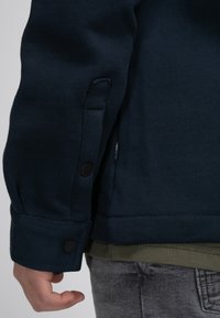 Marineblauwe sweatshirt met een gestructureerde stof, voorzien van een knoopdetail bij de mouwen en een gelaagde olijfgroene shirt zichtbaar eronder.