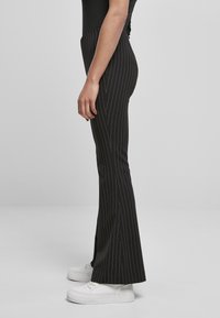 Sorte tapered bukser med vertikale hvite striper, designet med en utsvingt hem og brukt med hvite joggesko. Glatt stofftekstur.