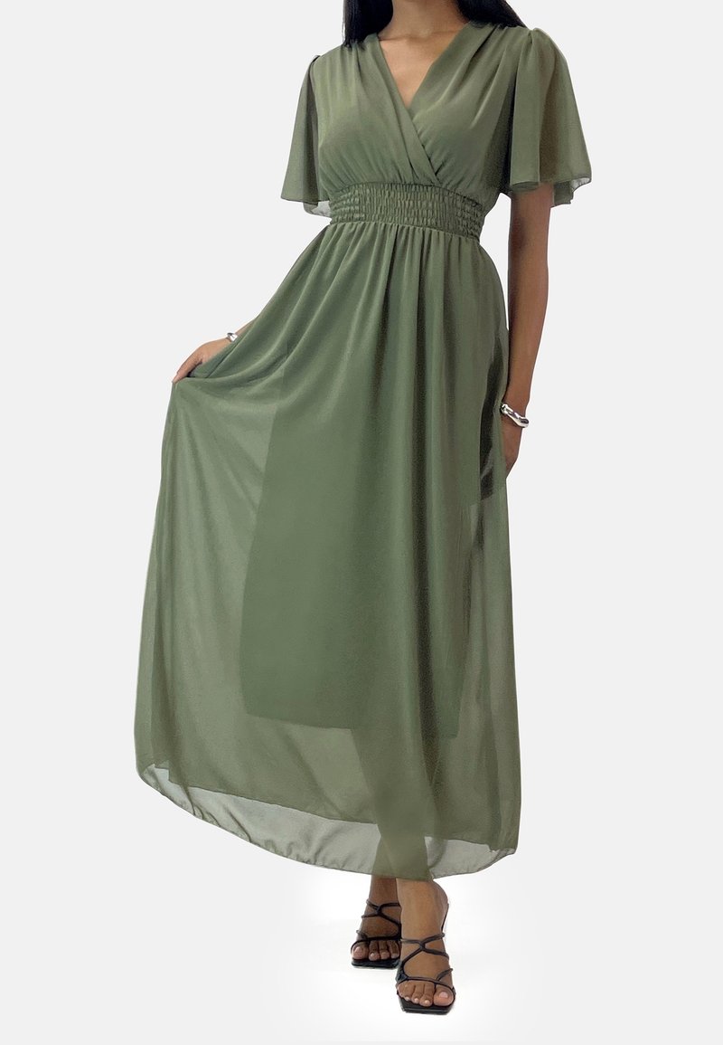 Femme portant une longue robe vert olive avec des manches volantes et des sandales noires à brides, tenant un côté de la robe.
