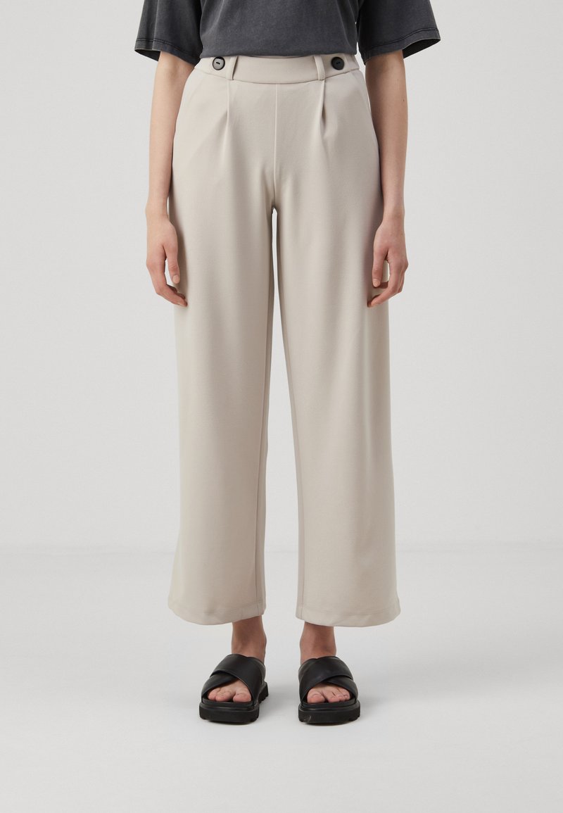 Pantalons beiges à jambes larges avec plis avant, deux boutons sur le côté et texture lisse. Associés à des sandales noires avec design à brides croisées.