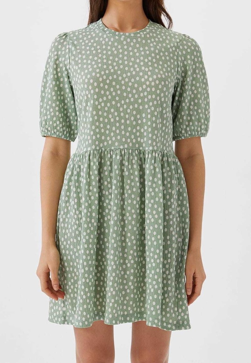 Femme portant une robe courte à manches courtes de couleur vert clair avec des pois blancs irréguliers et une taille froncée, debout sur un fond uni.