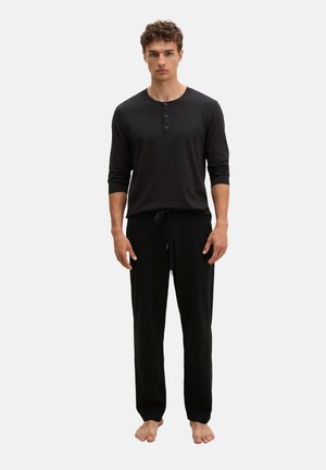 Junger Mann steht barfuß, trägt ein schwarzes langärmeliges Henley-Shirt und passende schwarze Kordelhose vor einem schlichten Hintergrund.