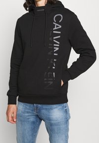 Černá šedá mikina s kapucí a logem ve tmavě šedé barvě na levé straně. Obsahuje šňůrky na stahování a klokankovou kapsu. Měkká tkanina, volný střih.