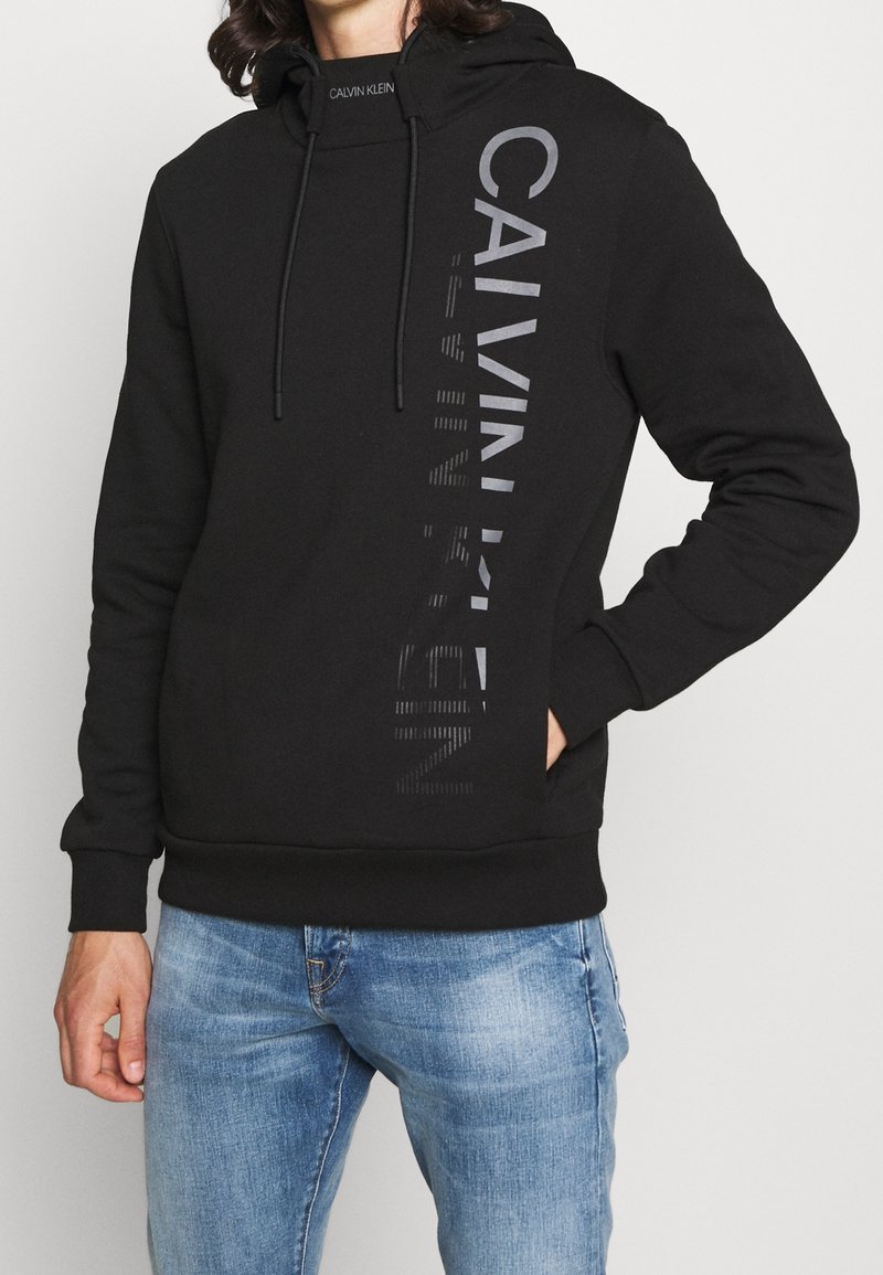 Černá šedá mikina s kapucí a logem ve tmavě šedé barvě na levé straně. Obsahuje šňůrky na stahování a klokankovou kapsu. Měkká tkanina, volný střih.