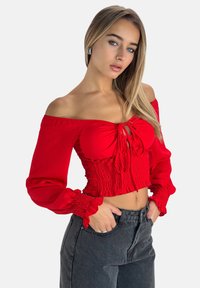 Blusa roja de hombros al aire con smocking elástico en la cintura y mangas abullonadas; cuenta con un detalle de lazo en el frente y una tela fluida.